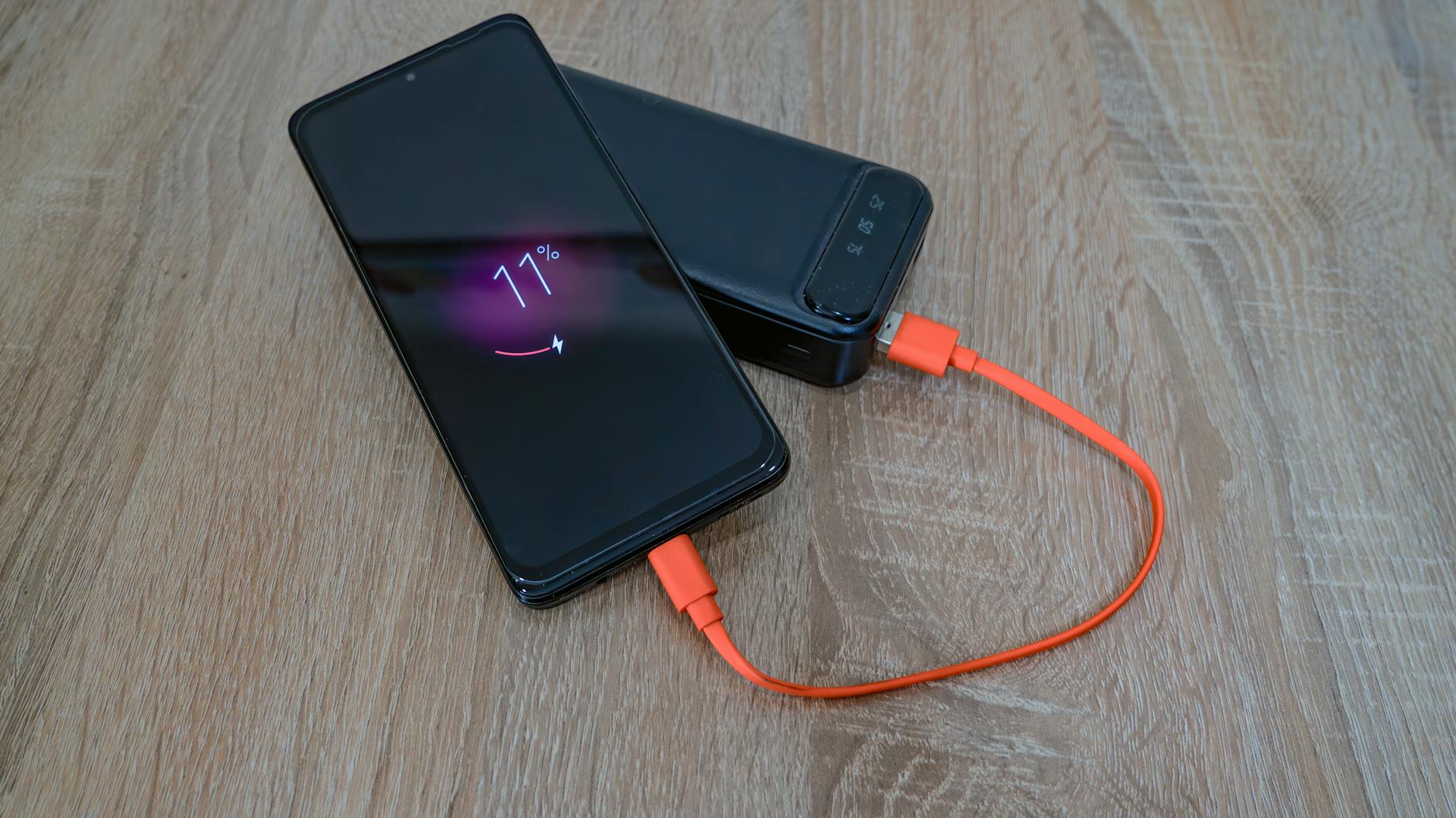 Smartfon ładowany z powerbanku pomarańczowym kablem na drewnianym stole